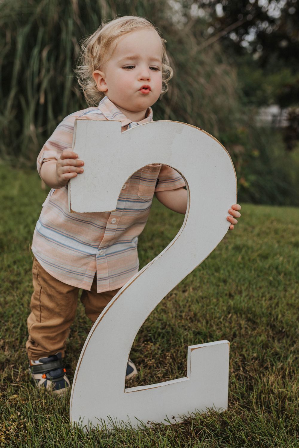 Toddler in striped shirt interacting with large white number two prop on lawn.