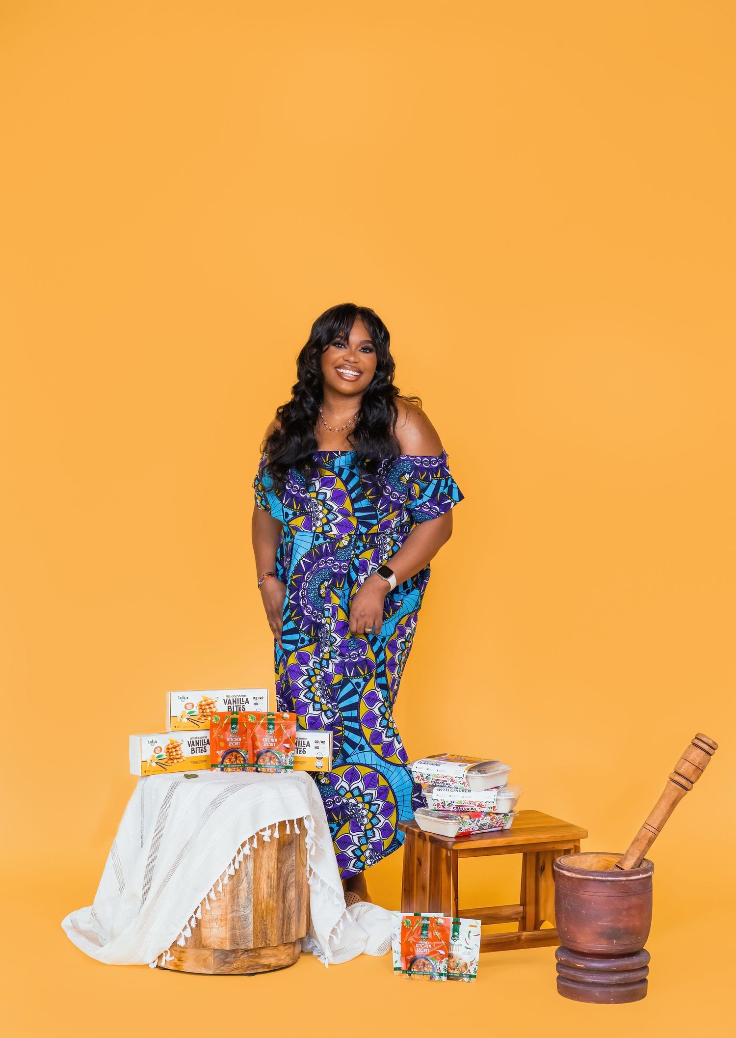 Woman in colorful floral dress poses with baking supplies against bright orange background.