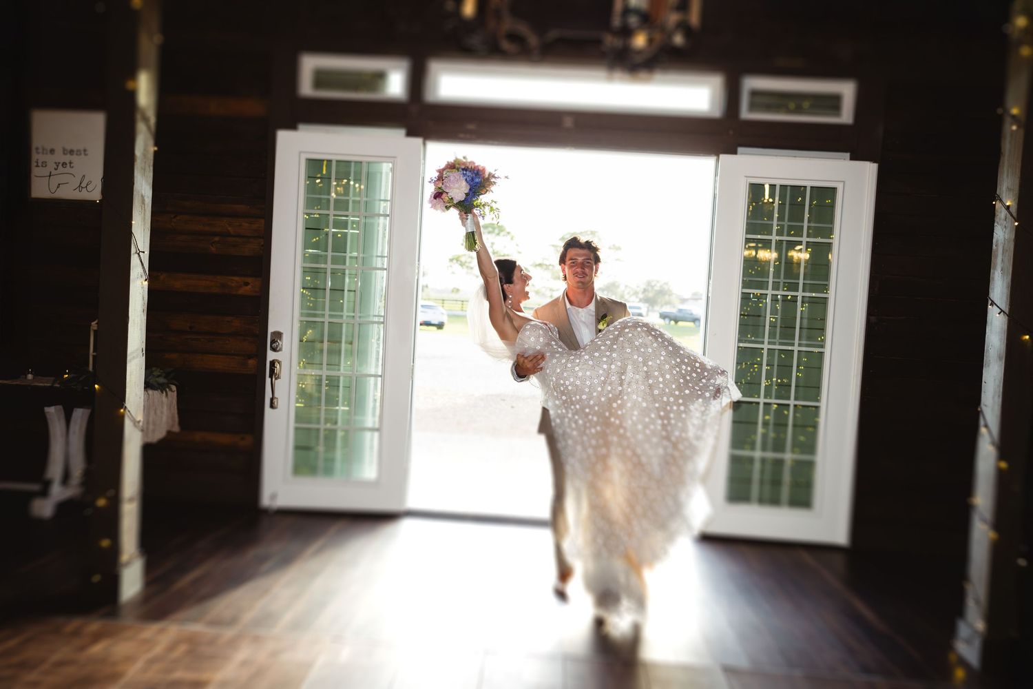 A joyful person in a flowing white lace wedding gown runs through a rustic barn doorway while holding a bouquet.
