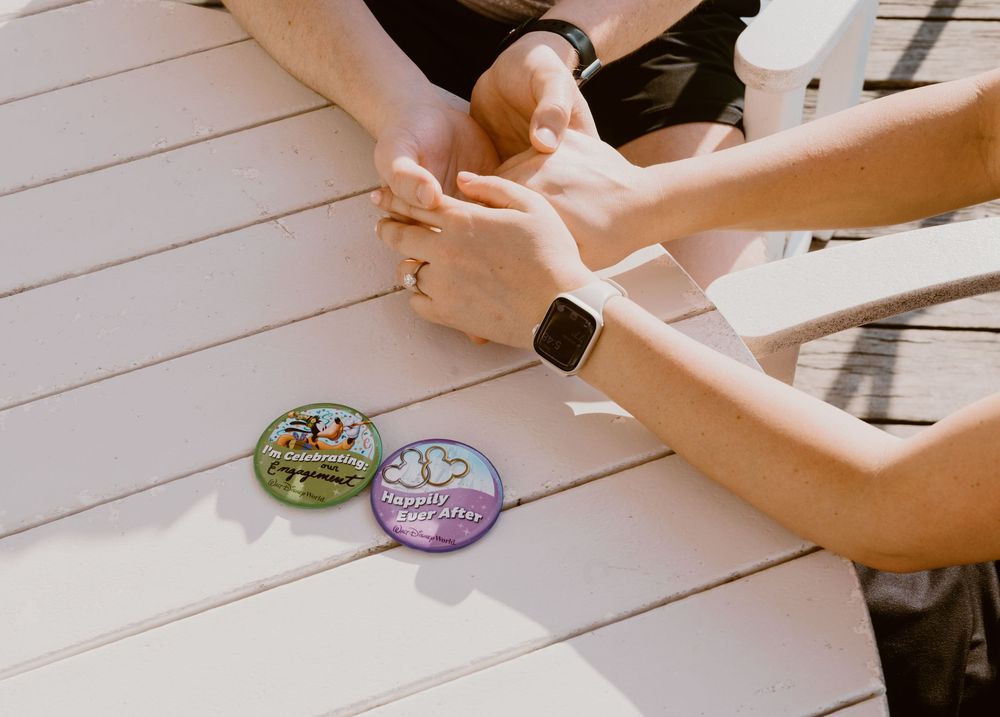 Young couple holding hands at sunset at Disney Boardwalk during surprise proposal with "I'm Celebrating" buttons.  Cred: LexTakePix