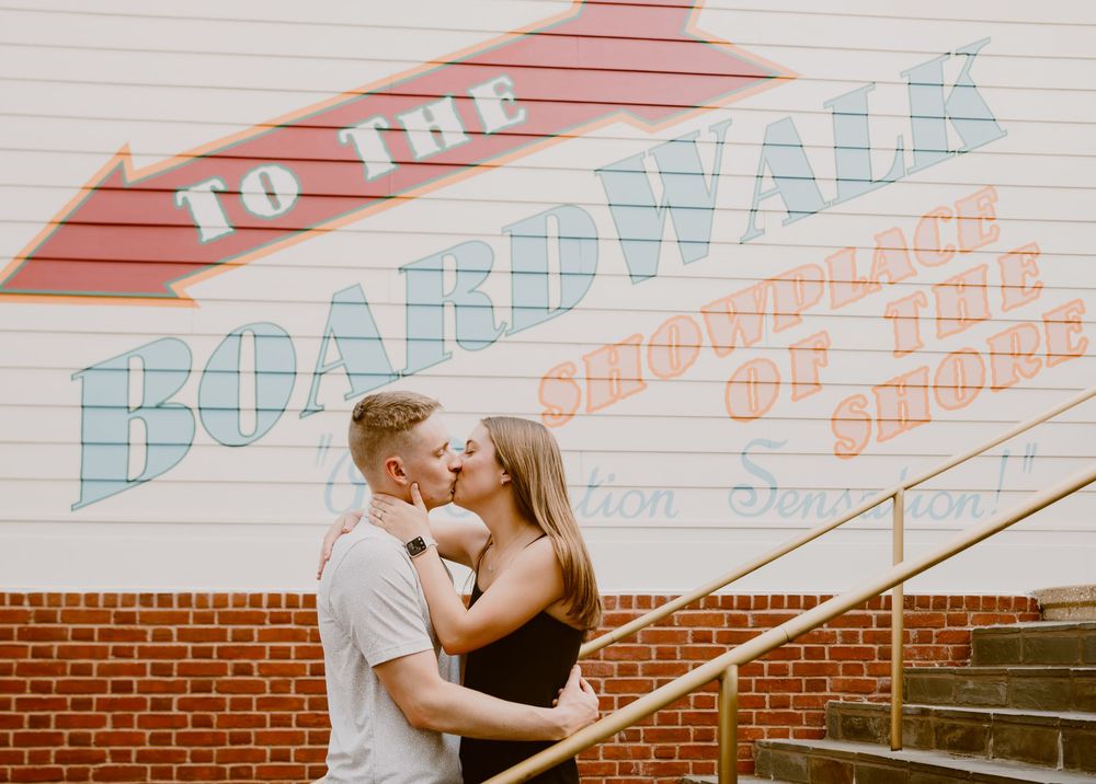 Young couple kissing at sunset at Disney Boardwalk during surprise proposal, at "To The Boardwalk" mural stairs at. Cred: LexTakePix
