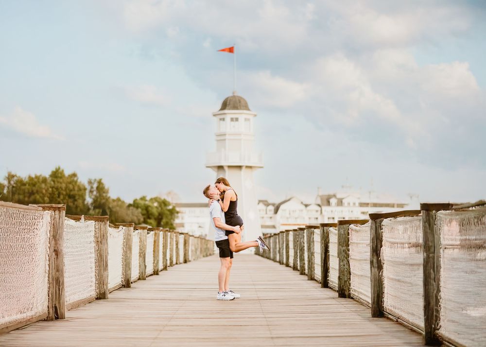 Young couple kissing at Disney Boardwalk in front of lighthouse during sunset at surprise proposal. Cred: LexTakePix