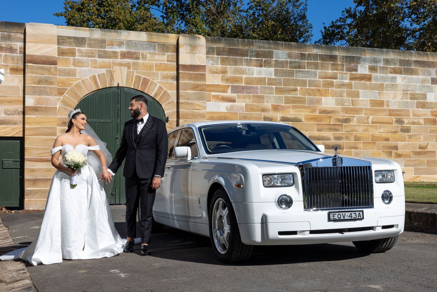 Wedding couple standing beside luxurious white Rolls-Royce against stone wall backdrop.
