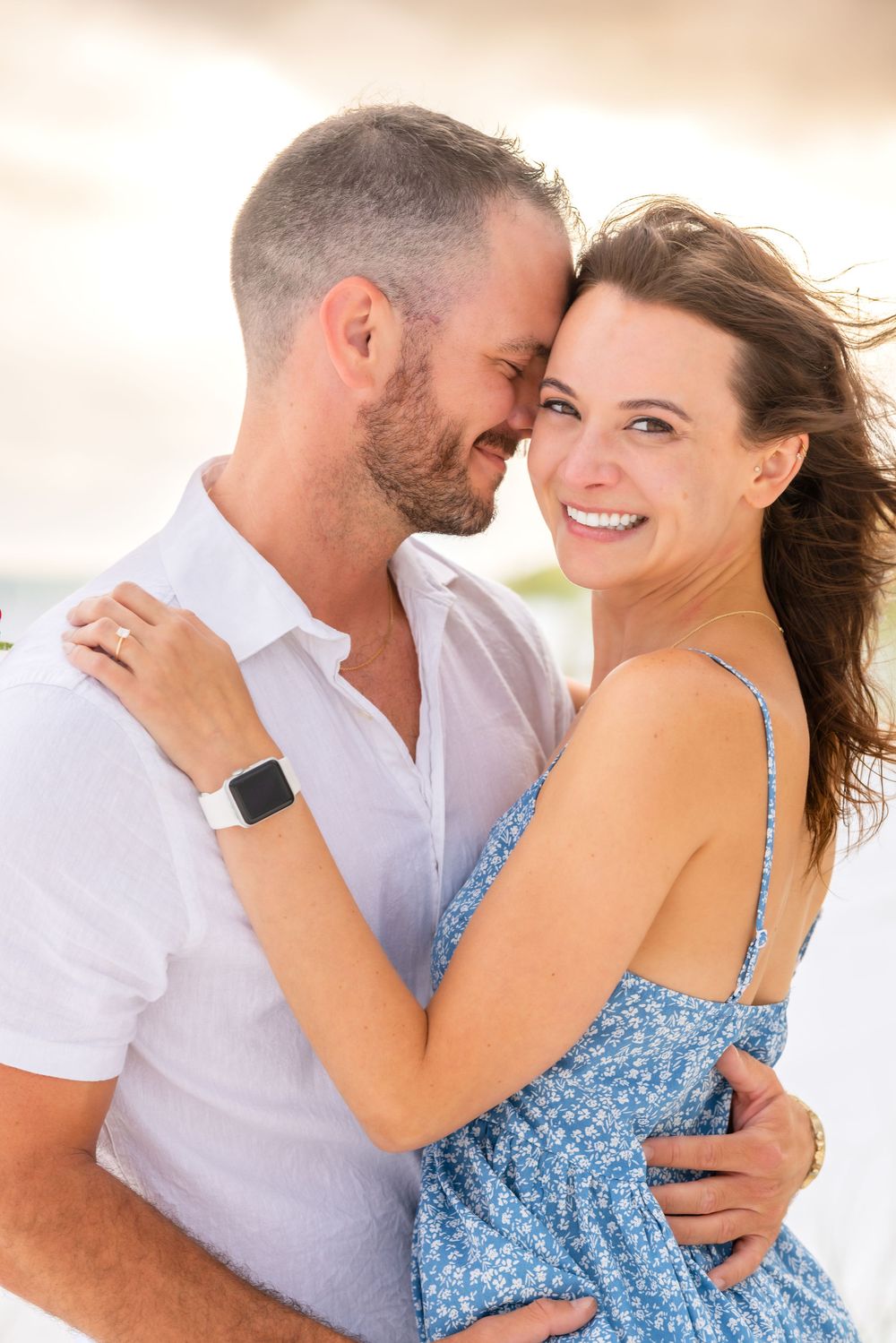 A loving couple embraces outdoors at sunset, with her wearing a blue floral dress and him in a white shirt.
