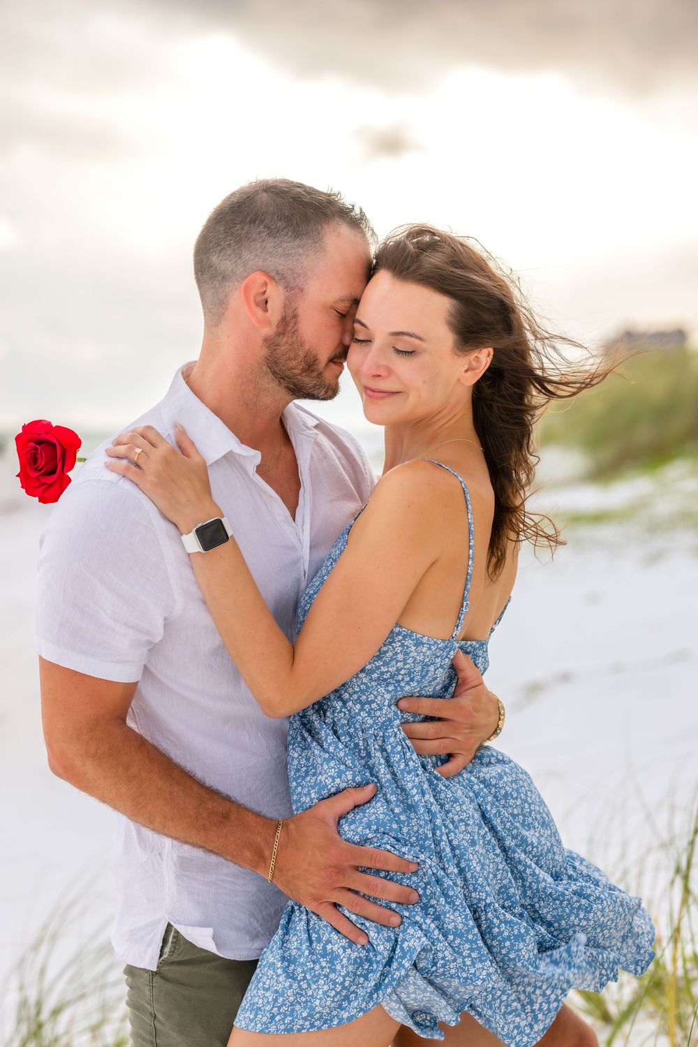A romantic couple embraces on a beach at sunset, with a red rose and the woman wearing a flowing blue floral dress.
