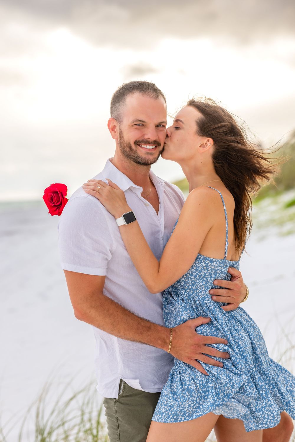 Romantic couple embraces on sandy beach with red rose as woman in blue dress leans in for a kiss at sunset.