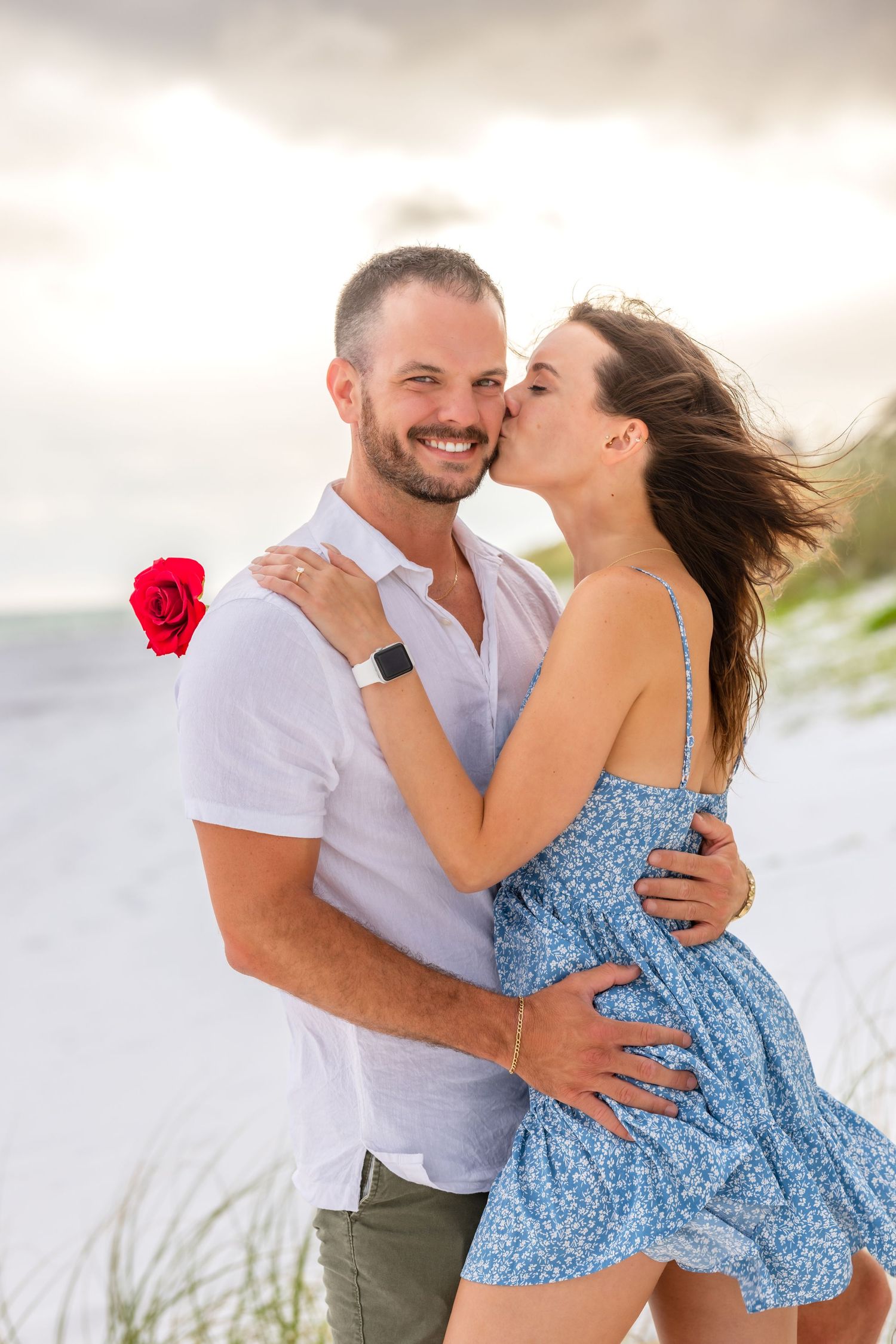 Romantic couple embraces on sandy beach with red rose as woman in blue dress leans in for a kiss at sunset.