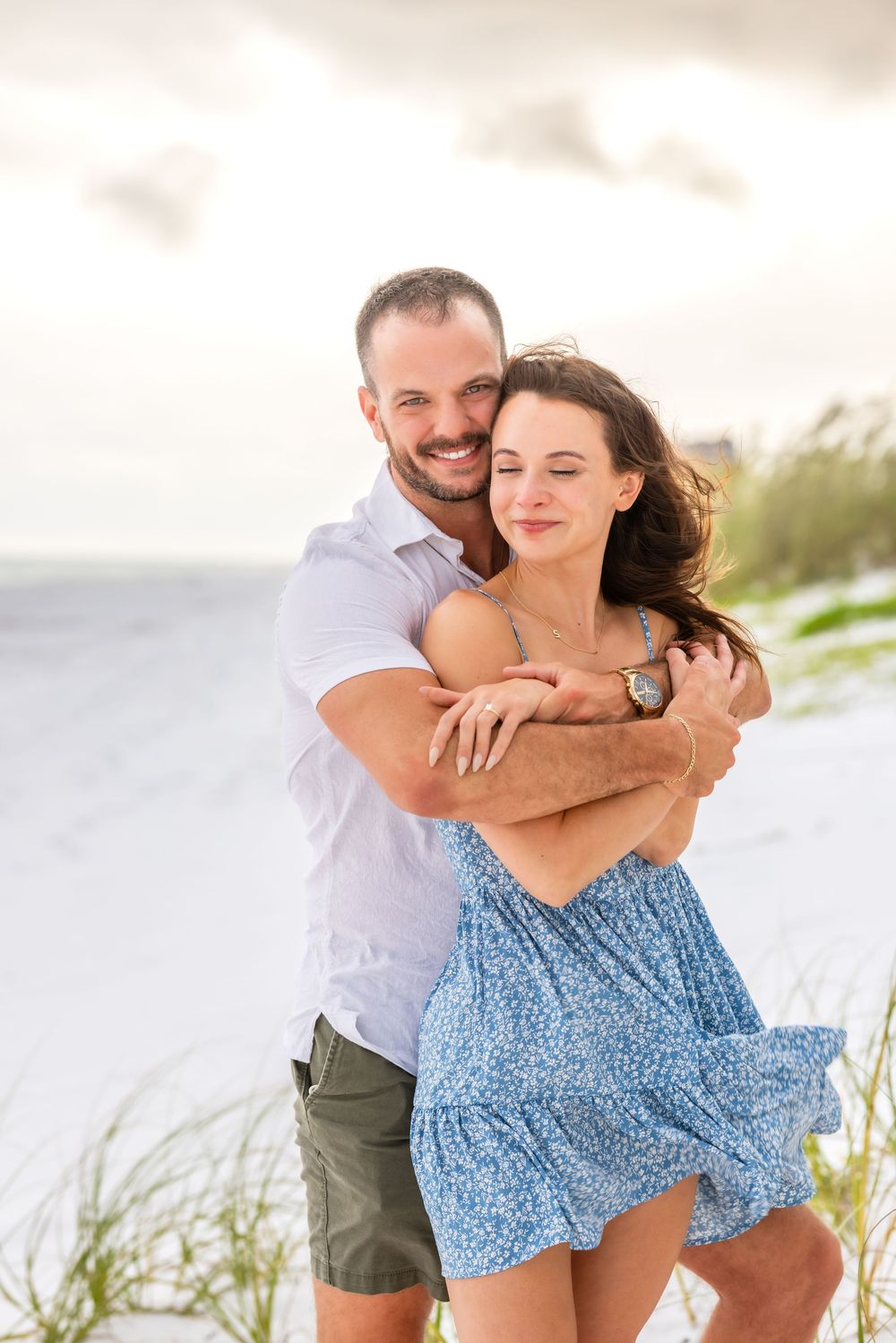 A romantic embrace on a sunny beach with soft sand and beach grass in the background.