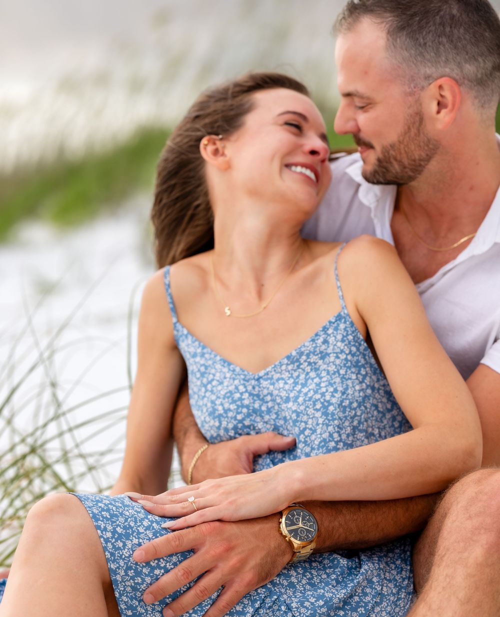 Couple embraces intimately on beach with sand dunes, woman wearing blue floral sundress and man in white shirt.