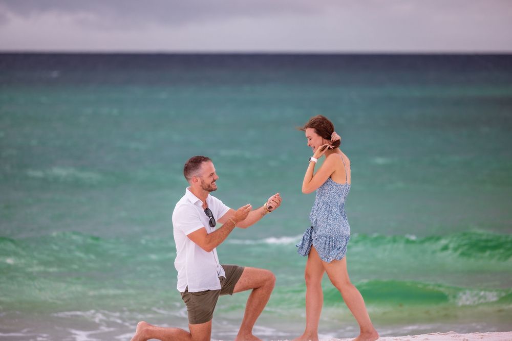A proposal moment unfolds on a beach with sparkling turquoise water in the background.