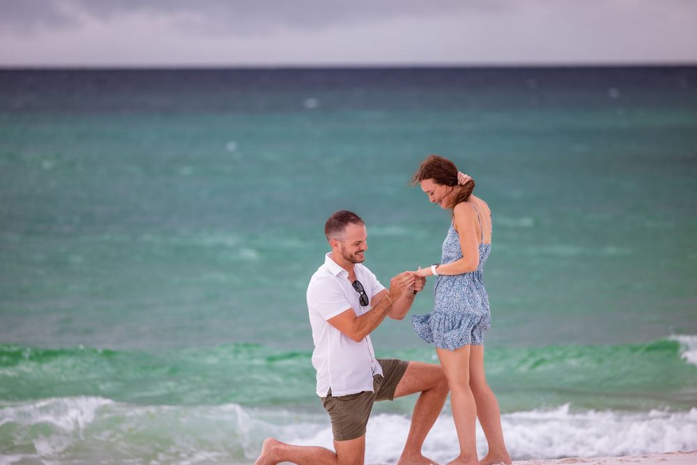 A romantic beach proposal moment captured on sandy shore with turquoise ocean waves in background.