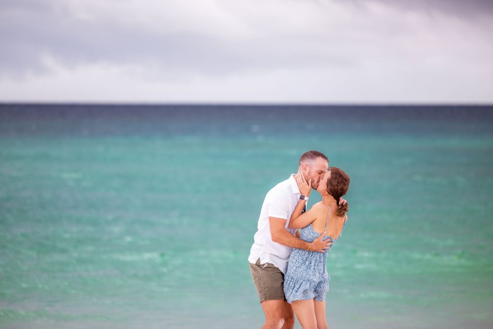 Couple embraces in a romantic kiss on the beach with turquoise waters and cloudy sky in the background.