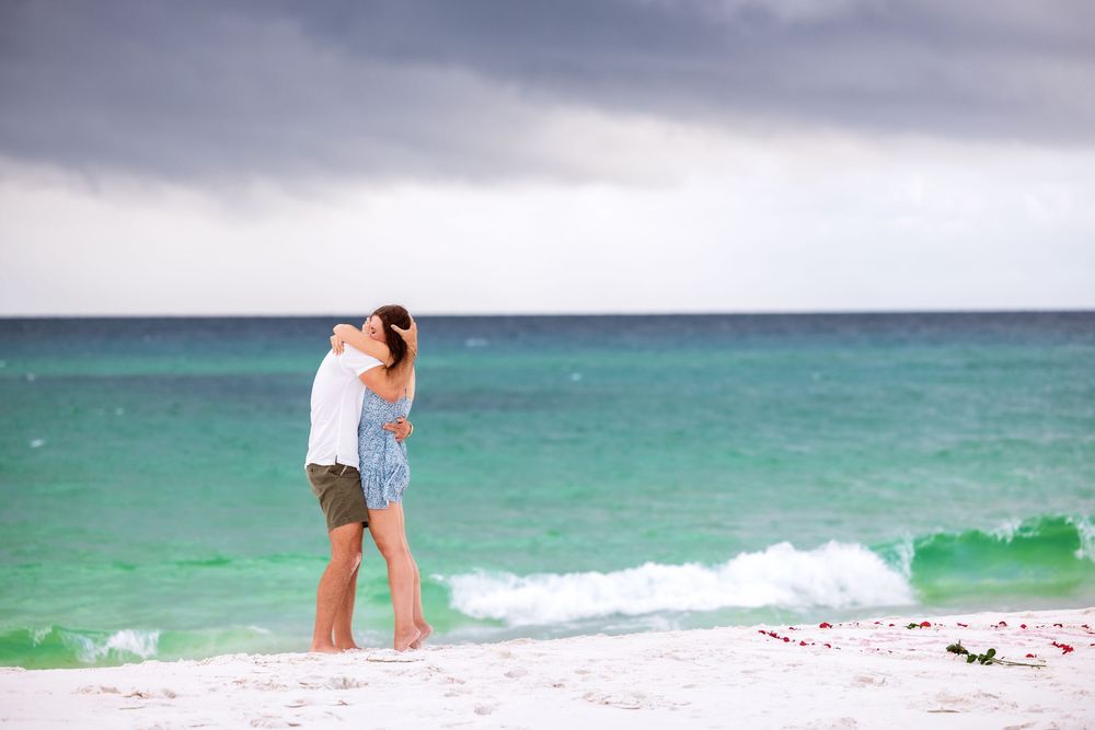 A couple embraces on a white sandy beach with turquoise waters and stormy gray clouds overhead.
