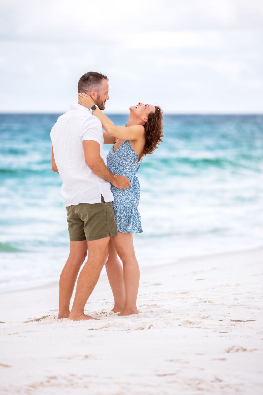 Couple embraces and laughs together on a sandy beach with turquoise ocean waves in the background.