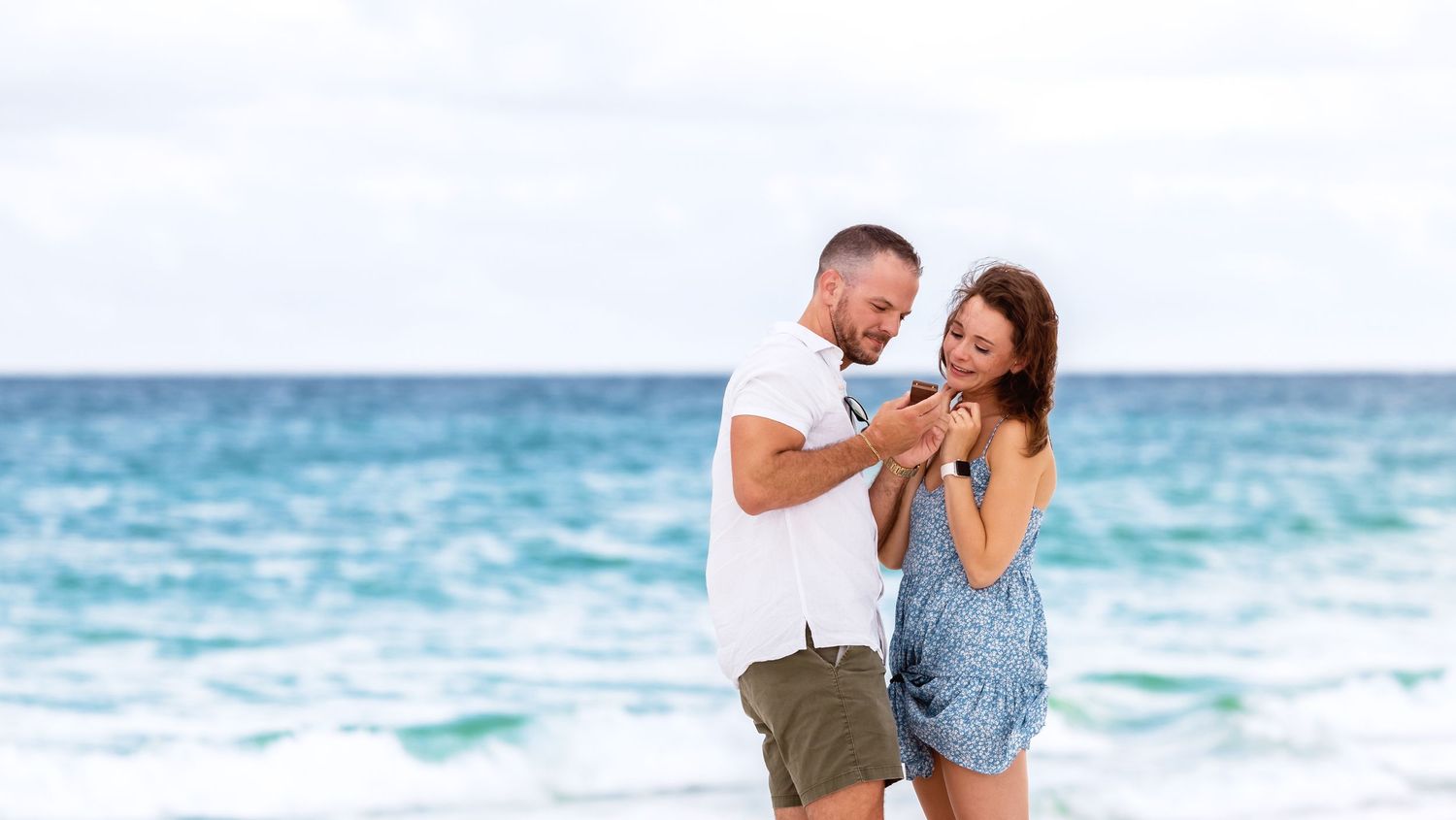 A couple shares a joyful moment together on a beach with turquoise ocean waves in the background.