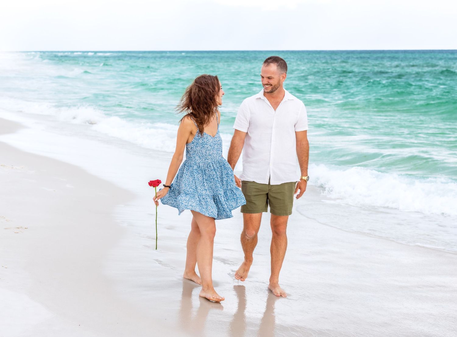 A couple walks hand in hand along a pristine beach with turquoise ocean waves in the background.