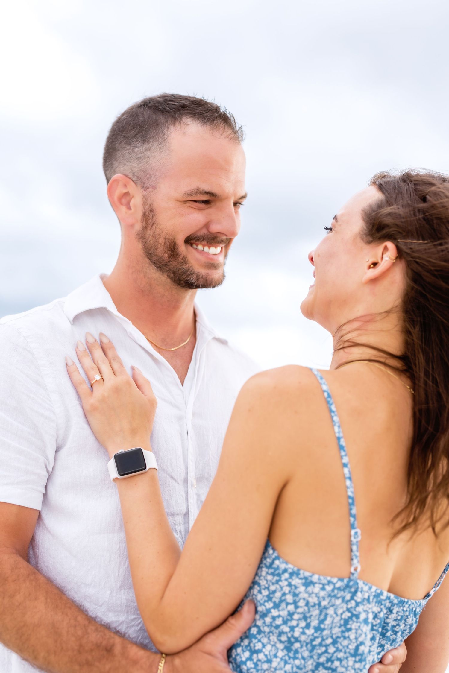A romantic couple in a blue dress and white shirt embrace happily during a photo shoot in natural light.
