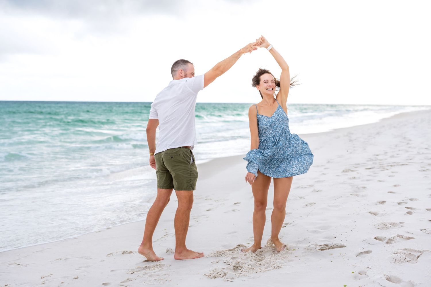 A couple dances together on a white sand beach with turquoise ocean waters in the background.