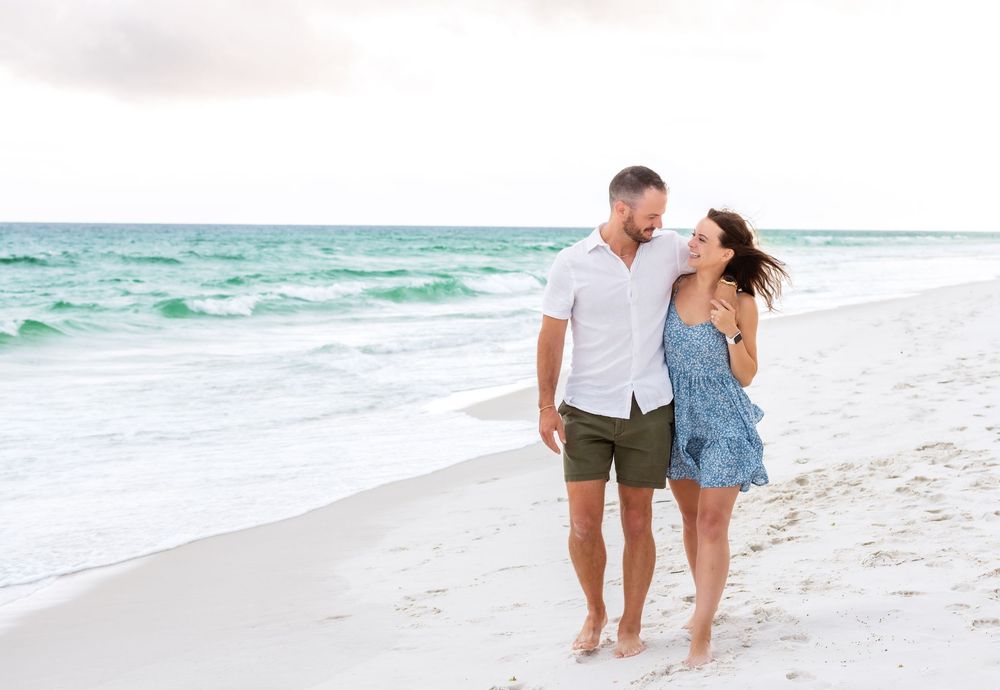 A couple walks along a white sandy beach with turquoise ocean waves in the background.