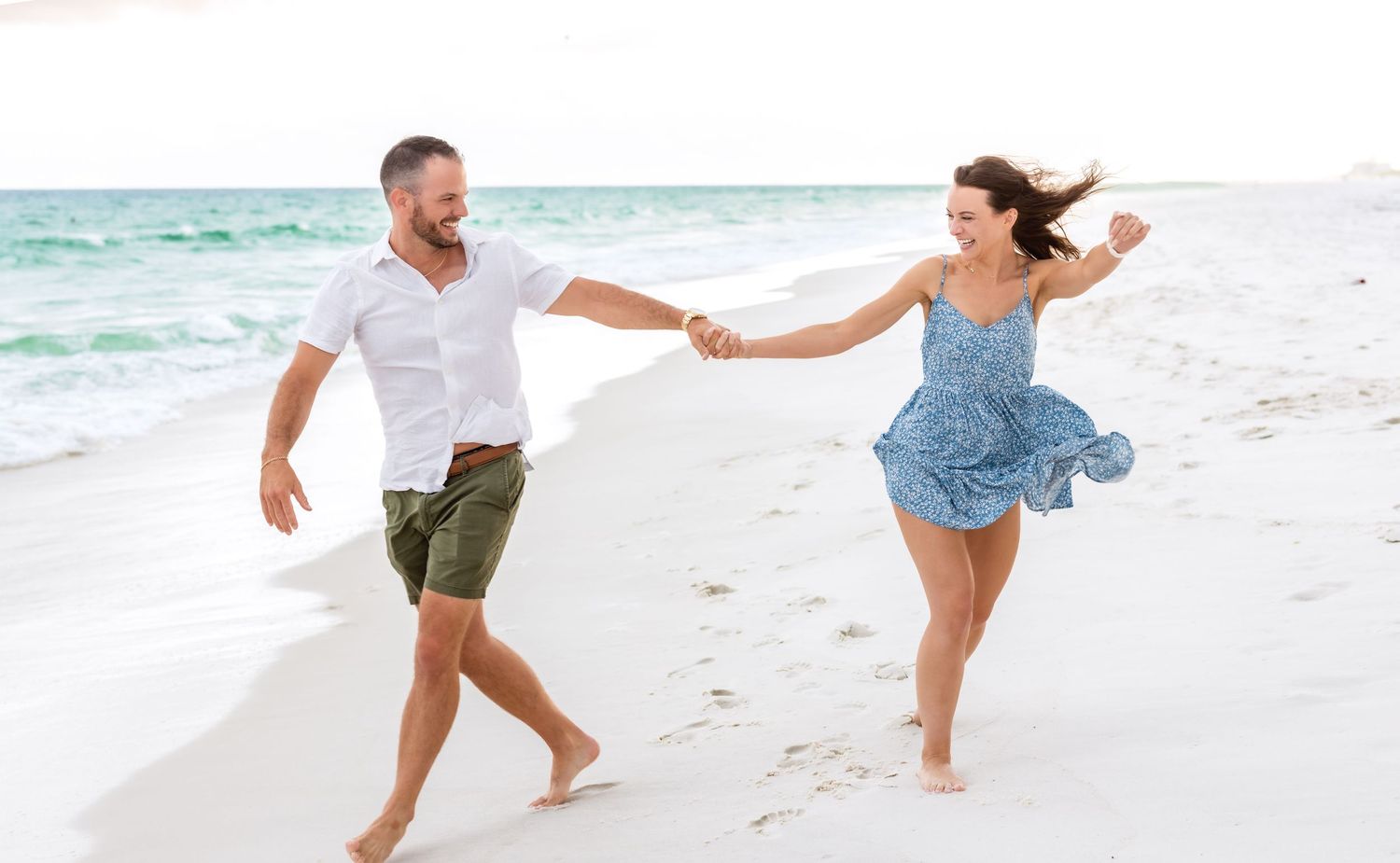 A happy couple runs and plays together along a pristine white sand beach with turquoise water in the background.