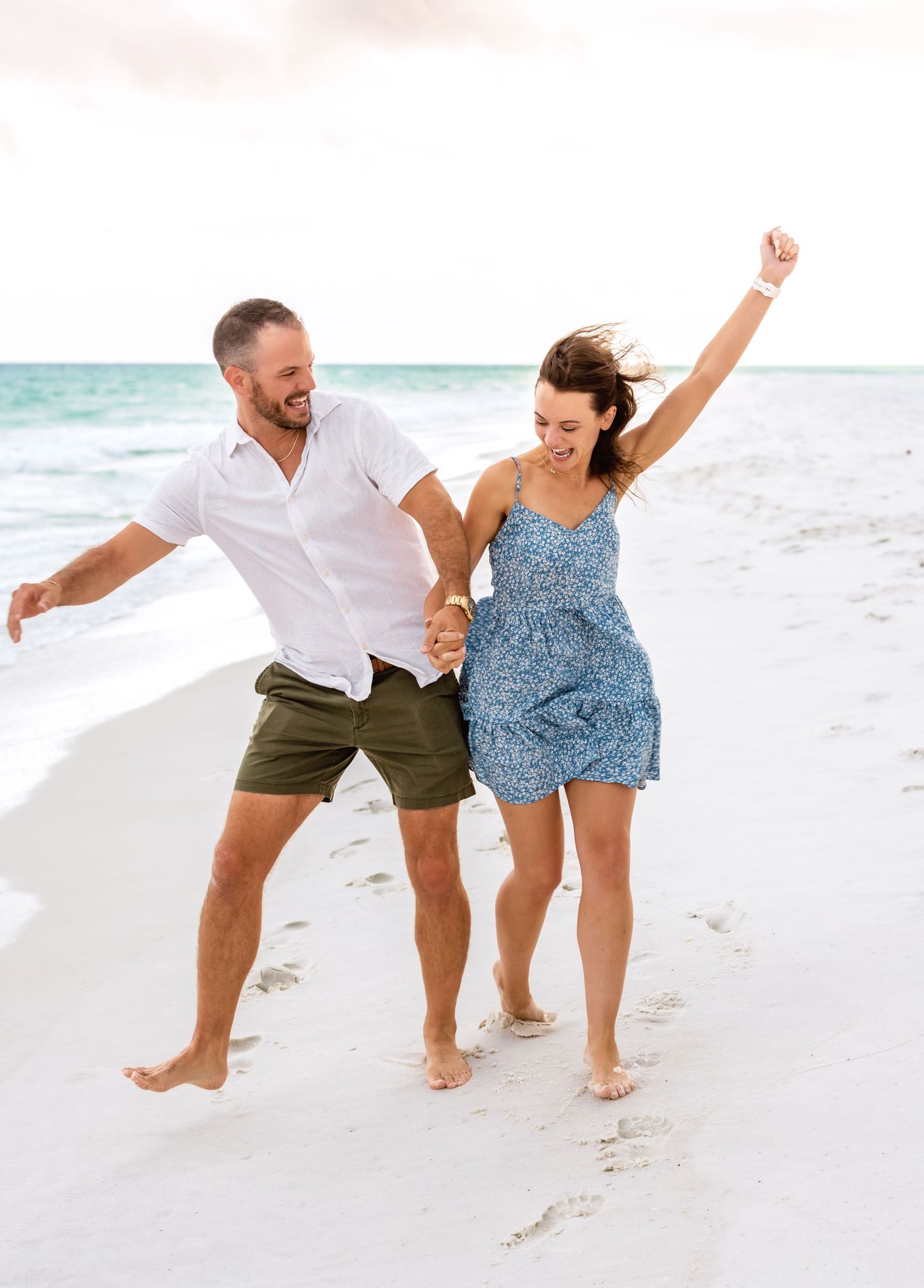 Couple walking playfully along a white sandy beach shore in casual summer outfits.