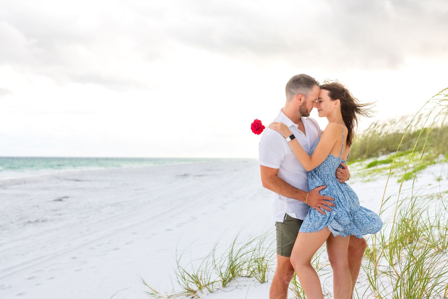 A romantic couple embraces on a peaceful beach with sea oats and ocean waves in the background.