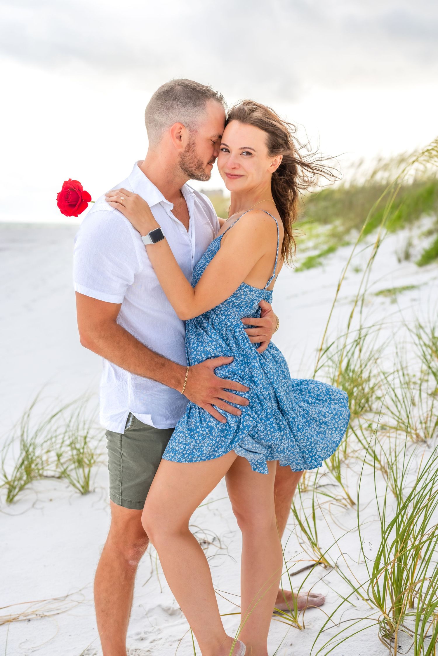 Romantic couple embraces on white sand beach with beach grass and red flower against cloudy sky.