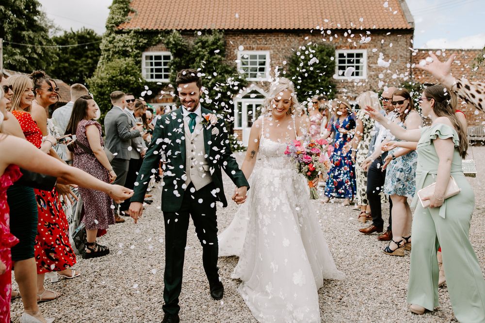 Wedding party celebration outside stone building with guests throwing white confetti along pathway.