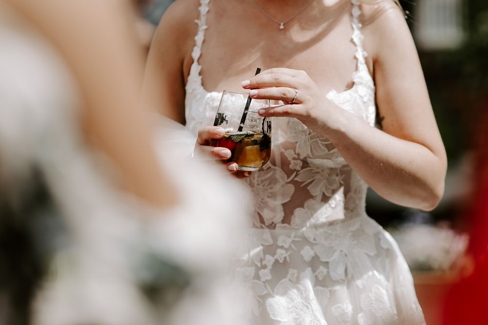 Someone in a white lace wedding dress holds a glass of whiskey or bourbon during a wedding celebration.