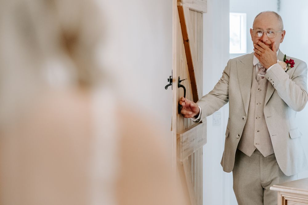Emotional first look reaction as someone wearing a beige suit and vest with red boutonniere sees their bride through doorway.