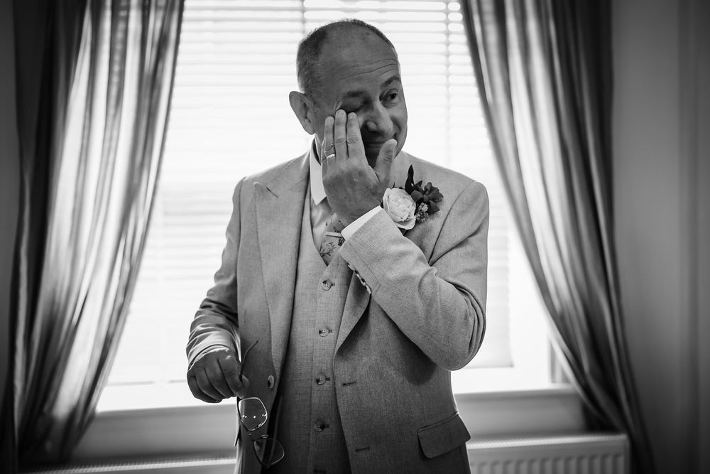 A groom in a gray suit adjusts his tie while standing next to curtained windows in black and white.