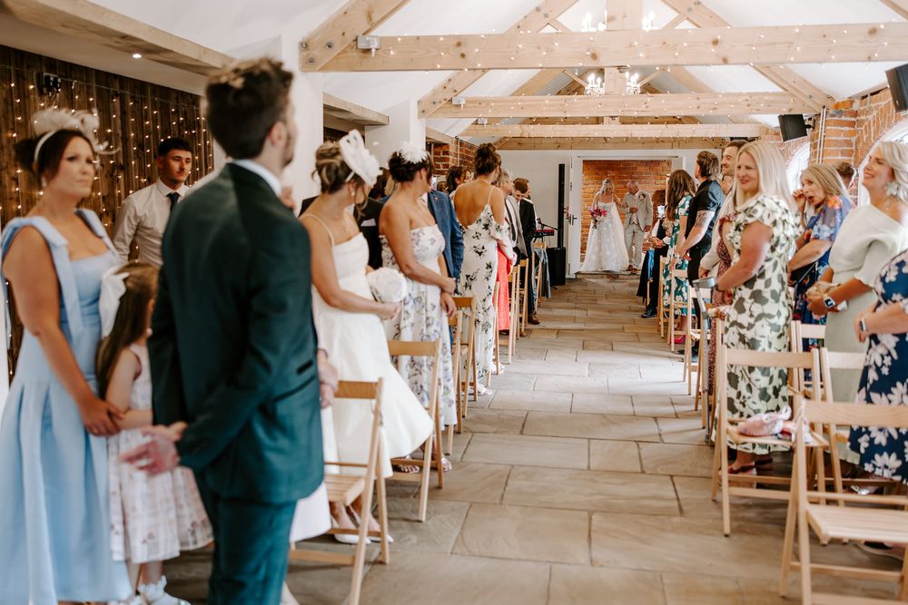 Wedding guests seated on wooden chairs in a rustic stone venue with exposed wooden beam ceiling and hanging chandeliers.