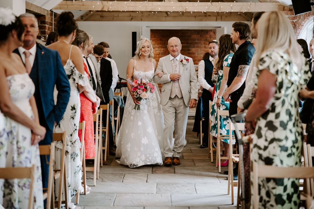 A couple walks down the aisle during a rustic barn wedding ceremony surrounded by guests in summer attire.