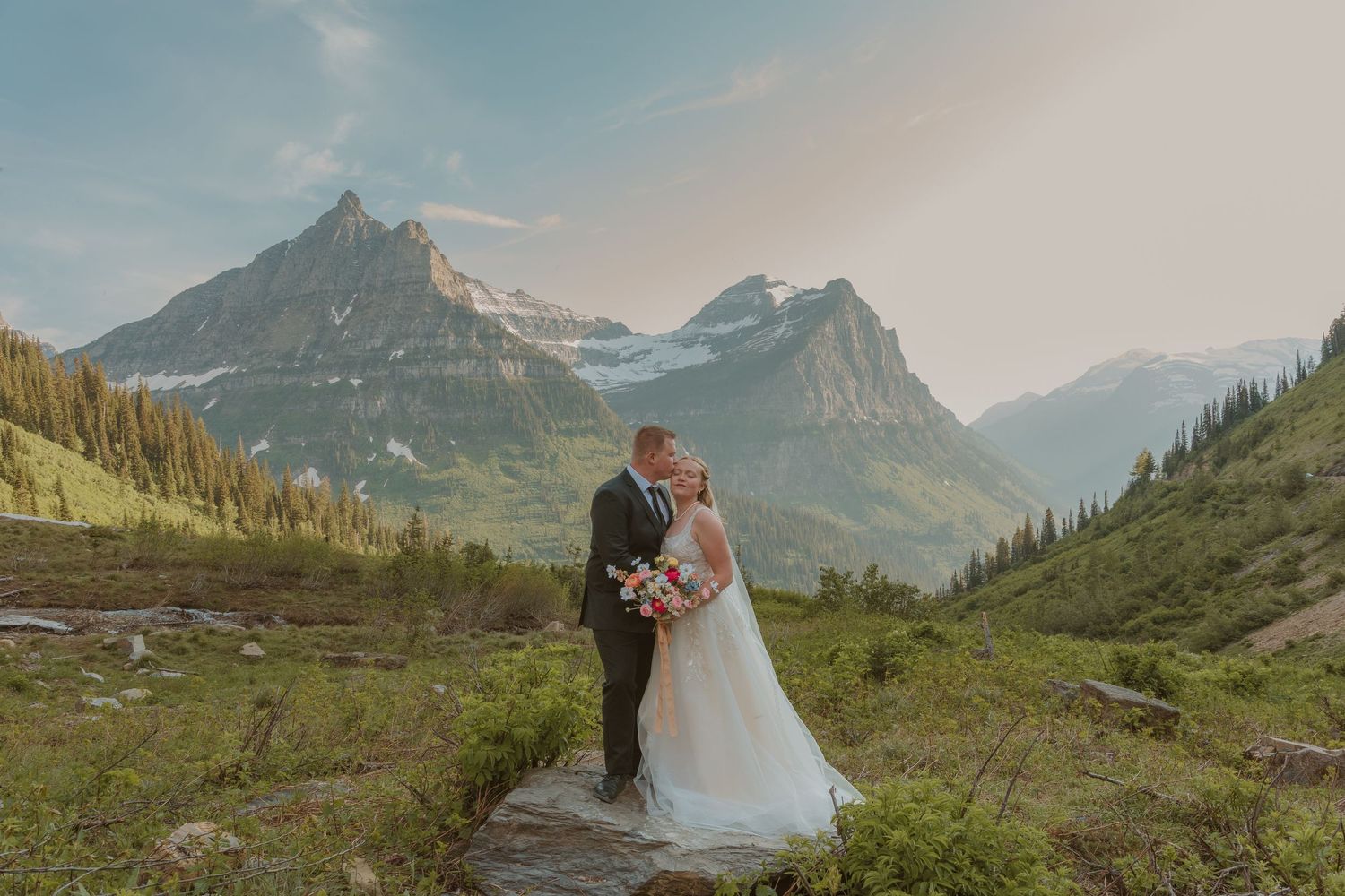A couple in wedding attire embrace on a scenic mountain overlook with dramatic peaks and evergreen forests in the background.