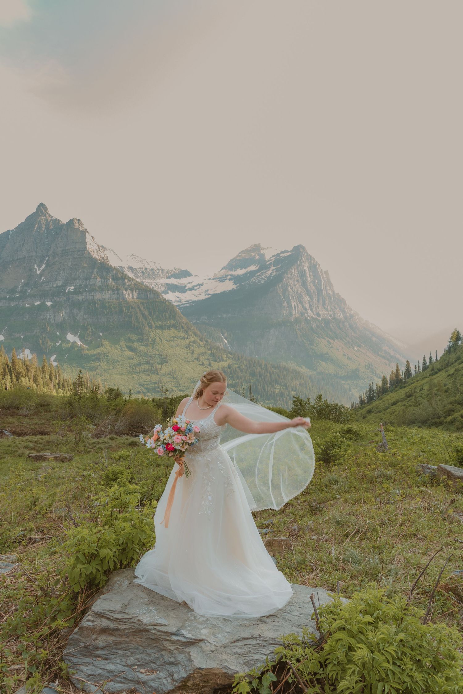 A flowing white wedding dress billows in the mountain breeze against a backdrop of majestic peaks in a natural landscape.