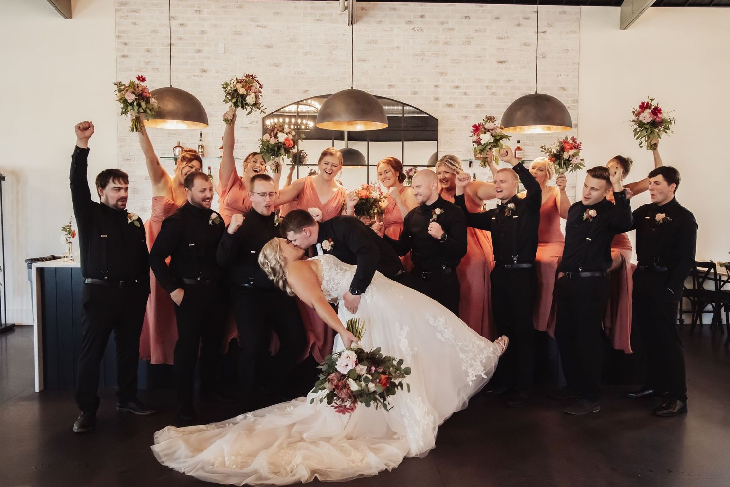 Wedding party in black and coral attire strikes a fun group pose during celebration at indoor venue.