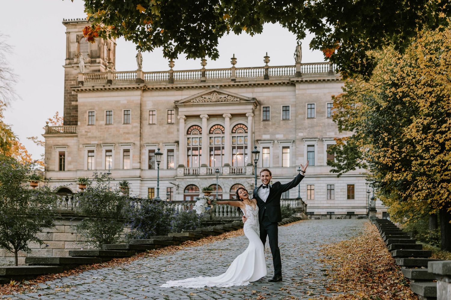 Couple celebrates their wedding day outside an elegant historic building with autumn trees framing the scene.