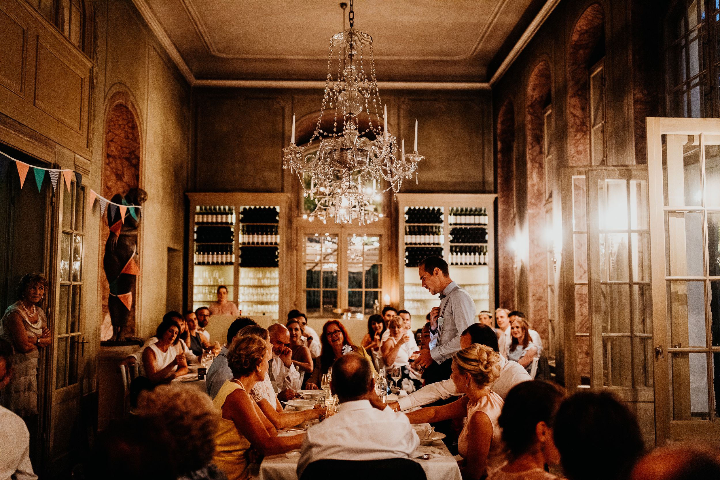 A candlelit dining room with crystal chandeliers shows guests seated at elegant tables during a formal dinner event.