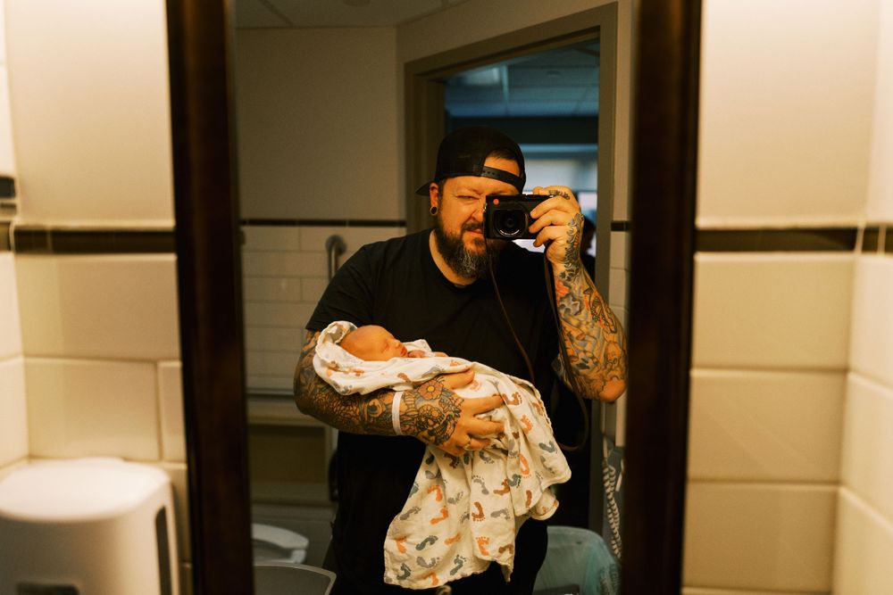 Person taking selfie in bathroom mirror while holding wrapped food items and wearing black clothing.