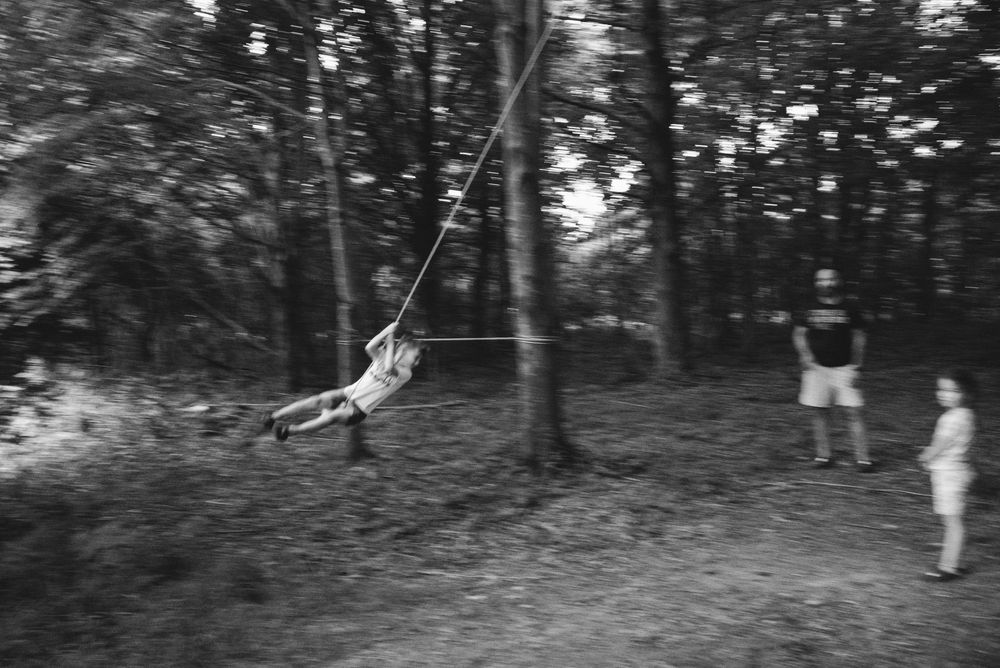 A blurred black and white photograph shows someone swinging on a rope swing in a wooded area while others watch.