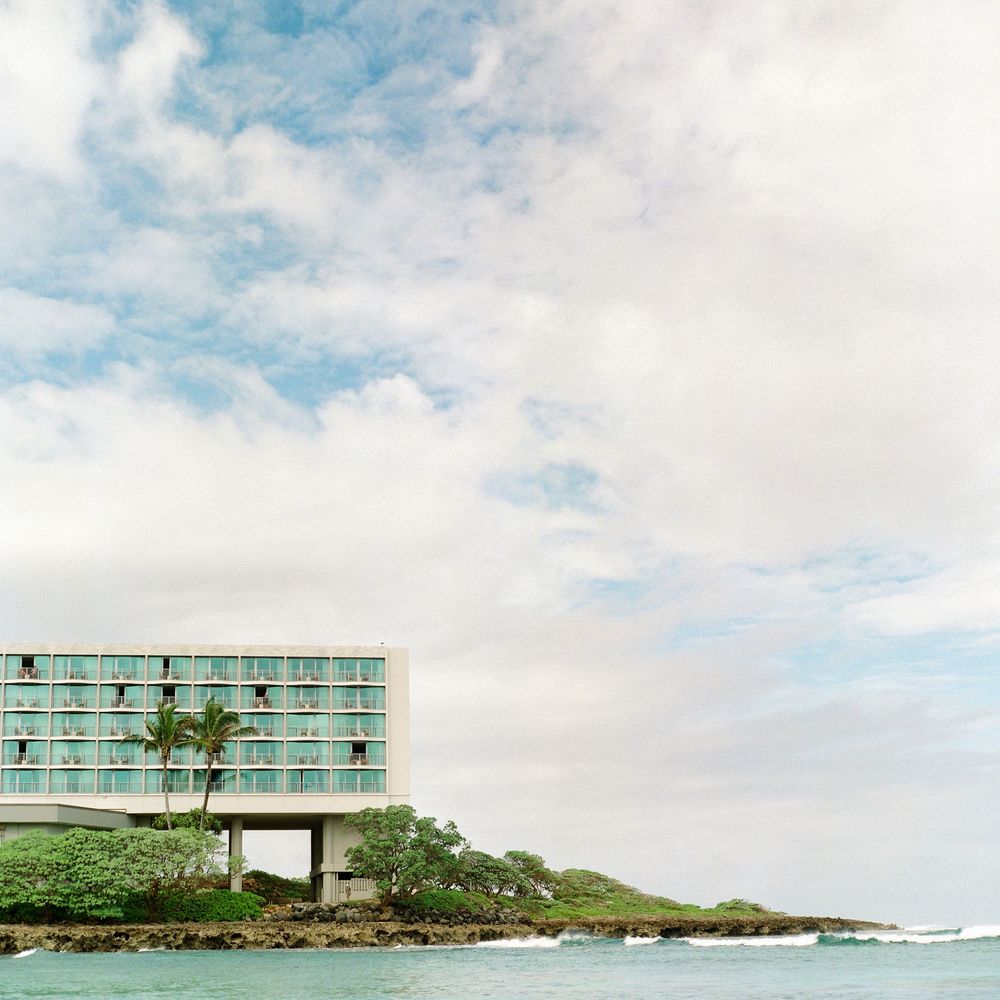 Panoramic view of a modern beachfront hotel building with palm trees and rocky coastline under cloudy skies.