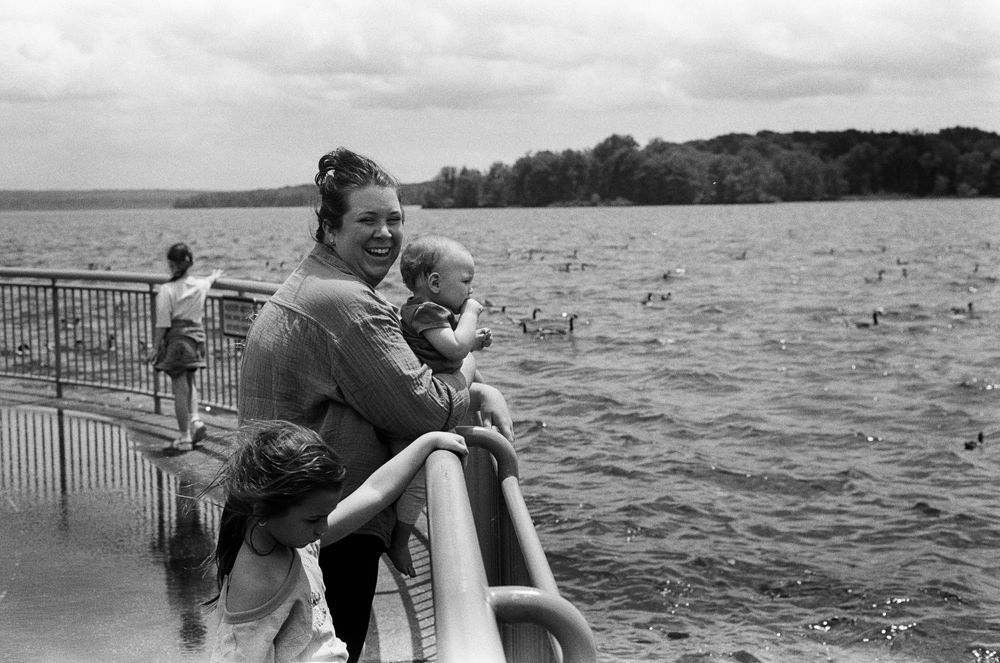 Black and white photo of a woman holding a baby on a lakeside railing, with trees and water in the background.