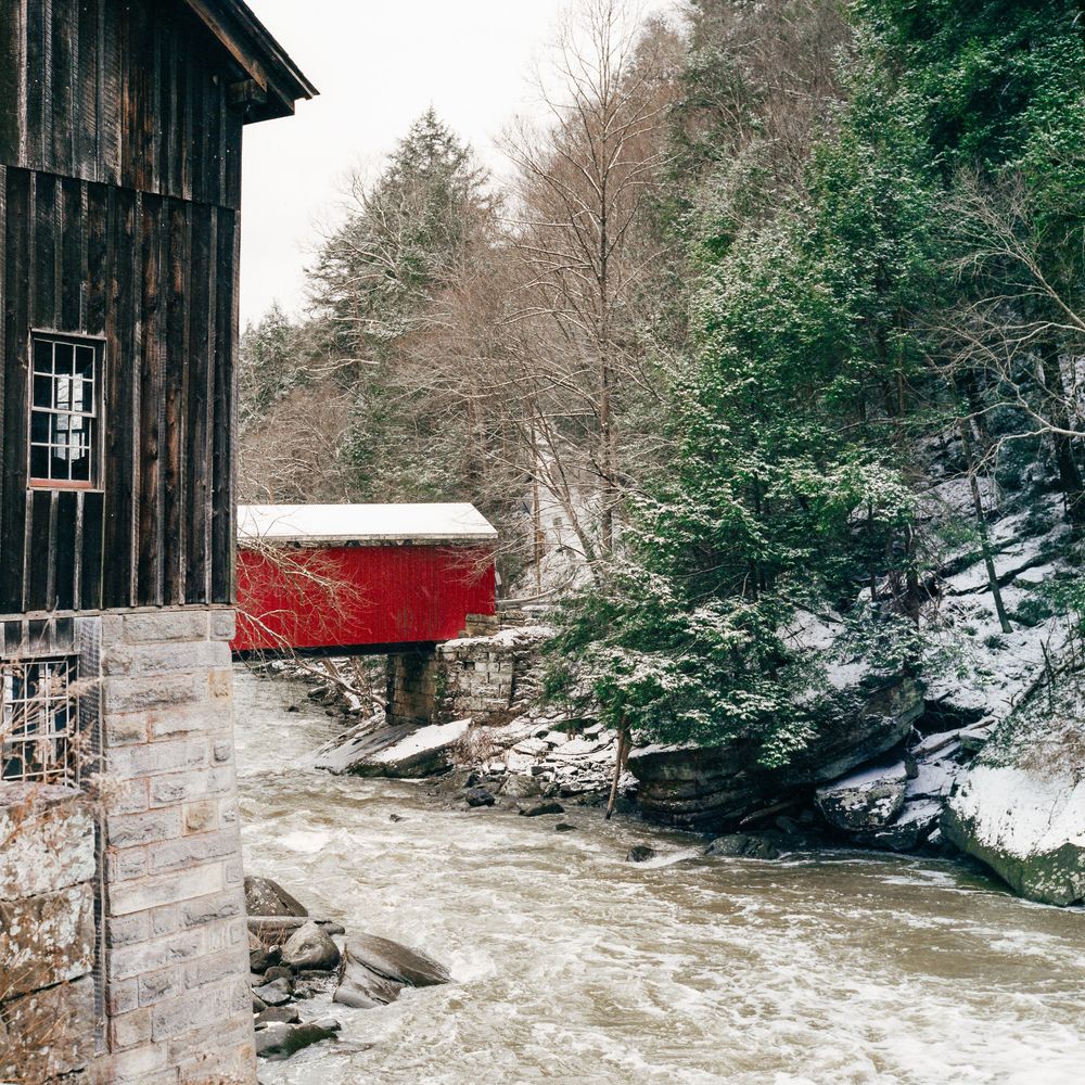 A snowy winter scene of a rushing river with a rustic wooden covered bridge visible in the distance.