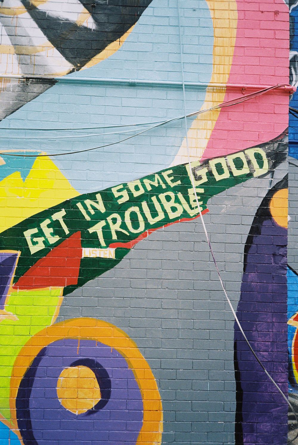 Colorful street art mural featuring the text 'Get in some good trouble' painted on a brick wall with abstract designs.