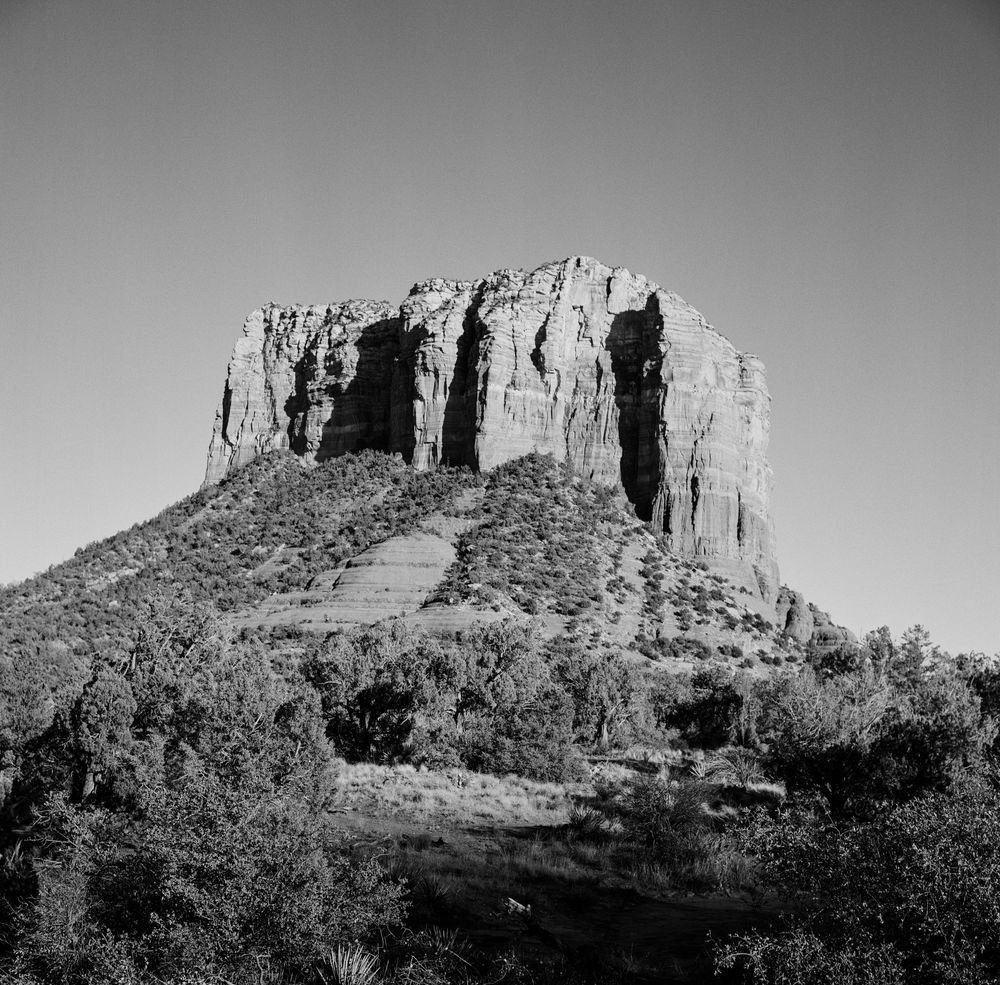 A dramatic black and white photograph of a towering butte formation in the desert with scrub vegetation below.