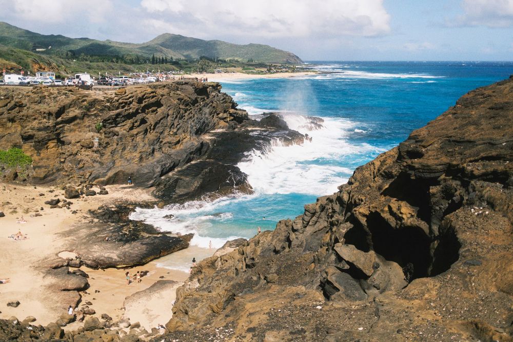 Rocky volcanic coastline with crashing blue waves against cliffs and sandy beach cove in Hawaii.
