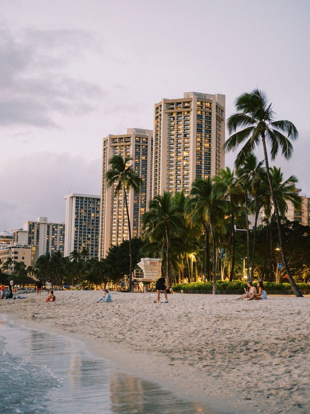 Tall beachfront hotels and swaying palm trees line Waikiki Beach at dusk as beachgoers enjoy the sandy shore.