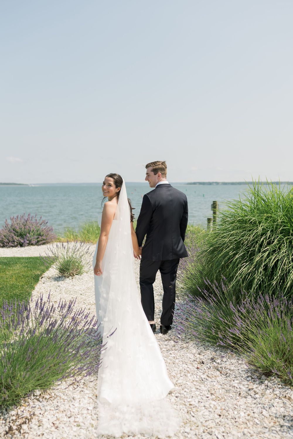 Coastal wedding portrait with couple walking along a lavender-lined path by the ocean.