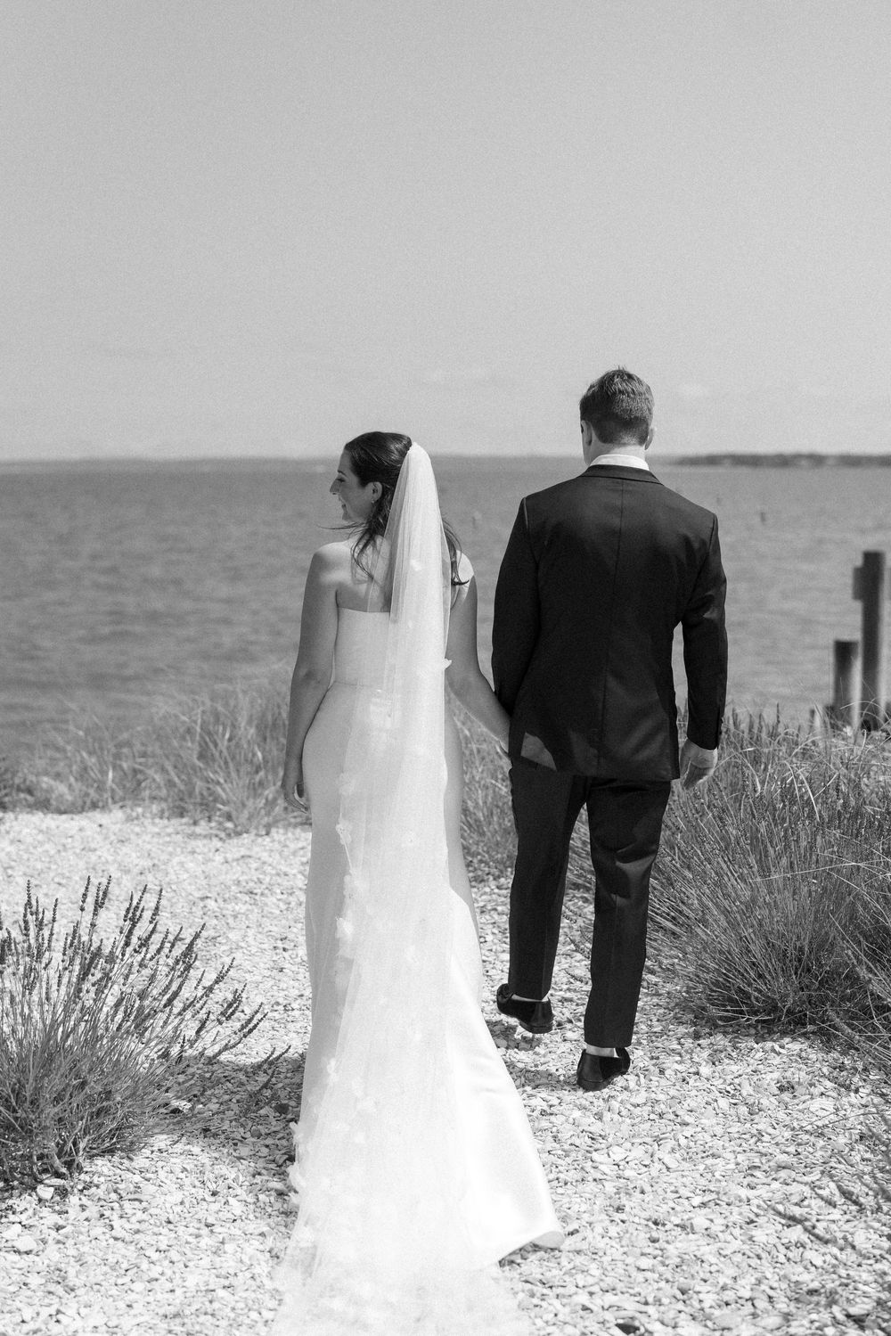 Black and white wedding photo of couple on coastal path with ocean backdrop.