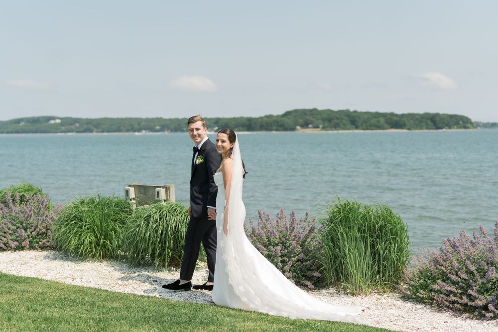 Waterfront wedding scene with couple embracing by lavender garden.